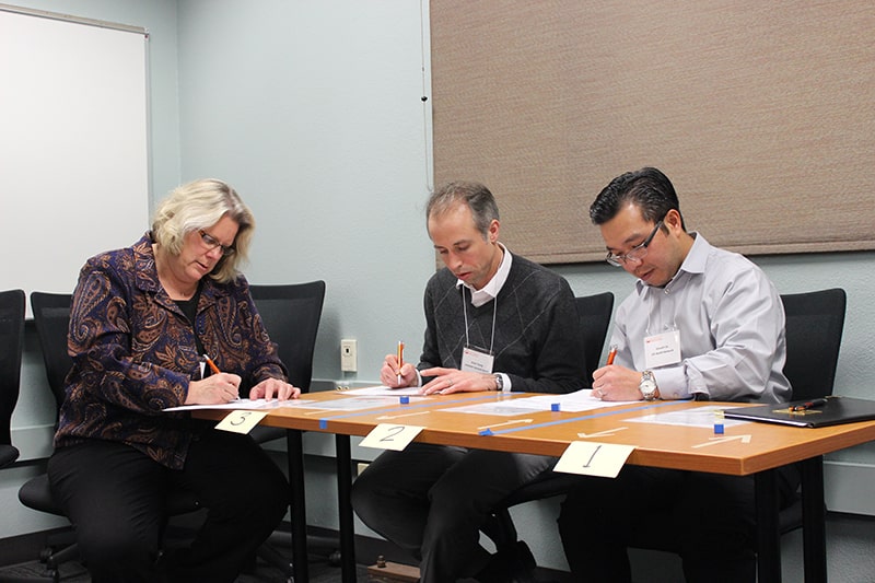 A panel of three instructors writing notes at a table