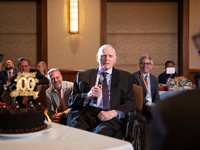 John Goodenough holds microphone at 100th birthday dinner with people smiling in the background